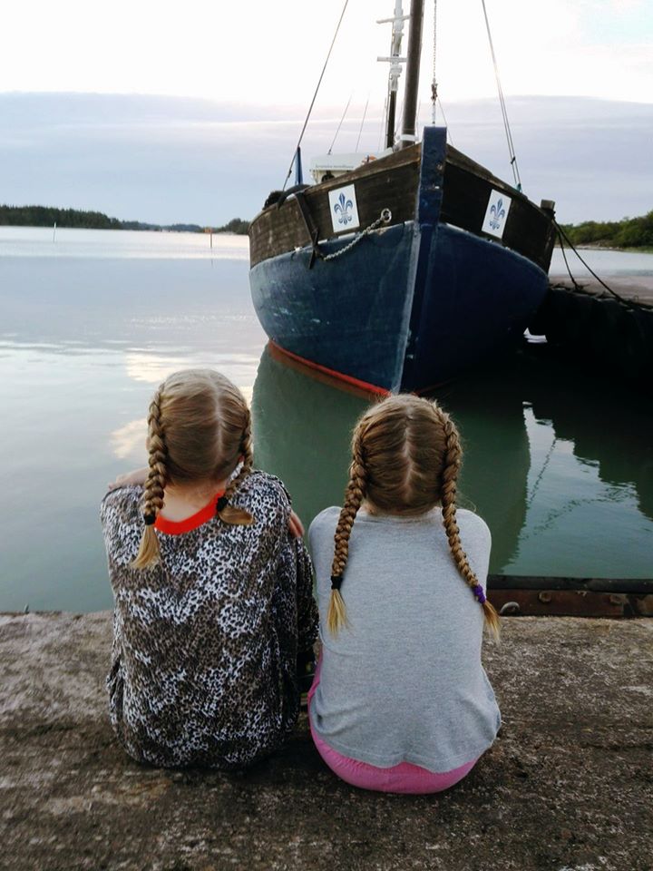 Kaksi tyttöä istuu rannalla ja katsovat laiturissa olevaa purjelaivaa