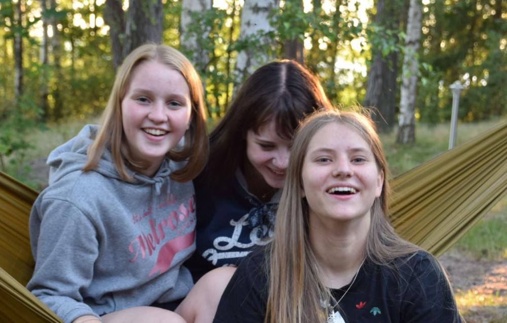 Kolme nuorta tyttöä istuu riippukeinussa riparilla ja hymyilee kameralle.